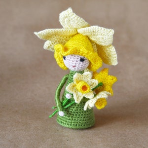 May include: A crocheted daffodil doll wearing a yellow flower hat and holding a bouquet of yellow daffodils.