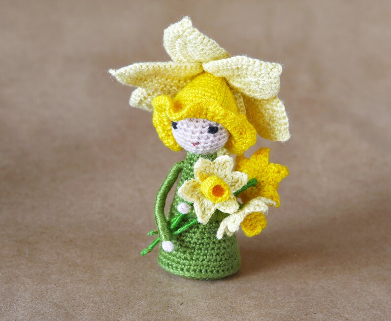 May include: A crocheted daffodil doll wearing a yellow flower hat and holding a bouquet of yellow daffodils.