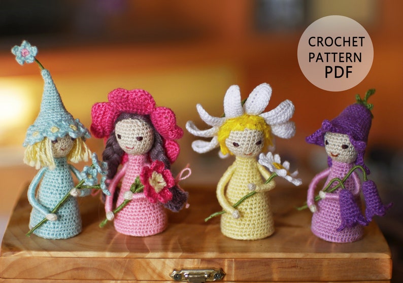 May include: Five crocheted dolls, each representing a different flower. The dolls are wearing colorful dresses and hats, and each holds a crocheted flower. The dolls are arranged in a row on a wooden surface. CROCHET PATTERN PDF