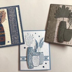 Puede incluir: Tres tarjetas de felicitación hechas a mano con un tema de jarrón y planta. La primera tarjeta tiene un jarrón azul con un patrón marrón y blanco y una planta marrón. La segunda tarjeta tiene un jarrón gris con una planta gris. La tercera tarjeta tiene un jarrón azul con una planta blanca.