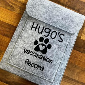 May include: A grey felt pet vaccination record holder with a flap. It displays "Hugo's" above a paw print with a heart, and the words "Vaccination Record" in black. The holder is on a wooden surface.