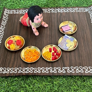 May include: A wooden tray with a white decorative border, featuring a small doll, four small bowls filled with various items, and a small plate with a note and a key. The bowls contain red and yellow sweets, red and yellow fruit, and a small amount of money.