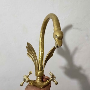May include: A gold-toned swan-shaped faucet with intricate details. The faucet has a curved spout and two handles. The swan's head is facing forward with its beak open.