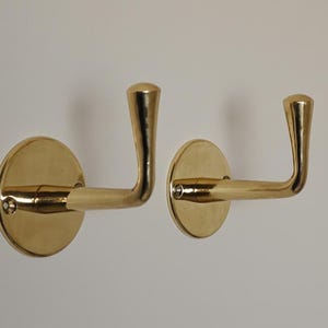 May include: Two brass wall hooks. Each hook has a circular base and a curved arm ending in a rounded tip. The hooks are mounted on a white wall, showcasing their polished, golden finish.