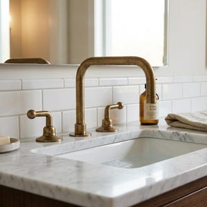 May include: A brass bathroom faucet with a high-arch spout and two lever handles mounted on a white marble countertop with a rectangular white sink basin. The faucet features a weathered metallic finish and is positioned against a white subway tile wall.