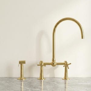 May include: A gold-toned kitchen faucet set with a high-arc spout, two cross-shaped handles, and a side sprayer. The set is displayed on a gray marble countertop against a cream-colored wall.