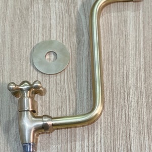 May include: A gold-toned faucet with a cross-shaped handle and a long, curved spout. The faucet is attached to a round, gold-toned wall plate.