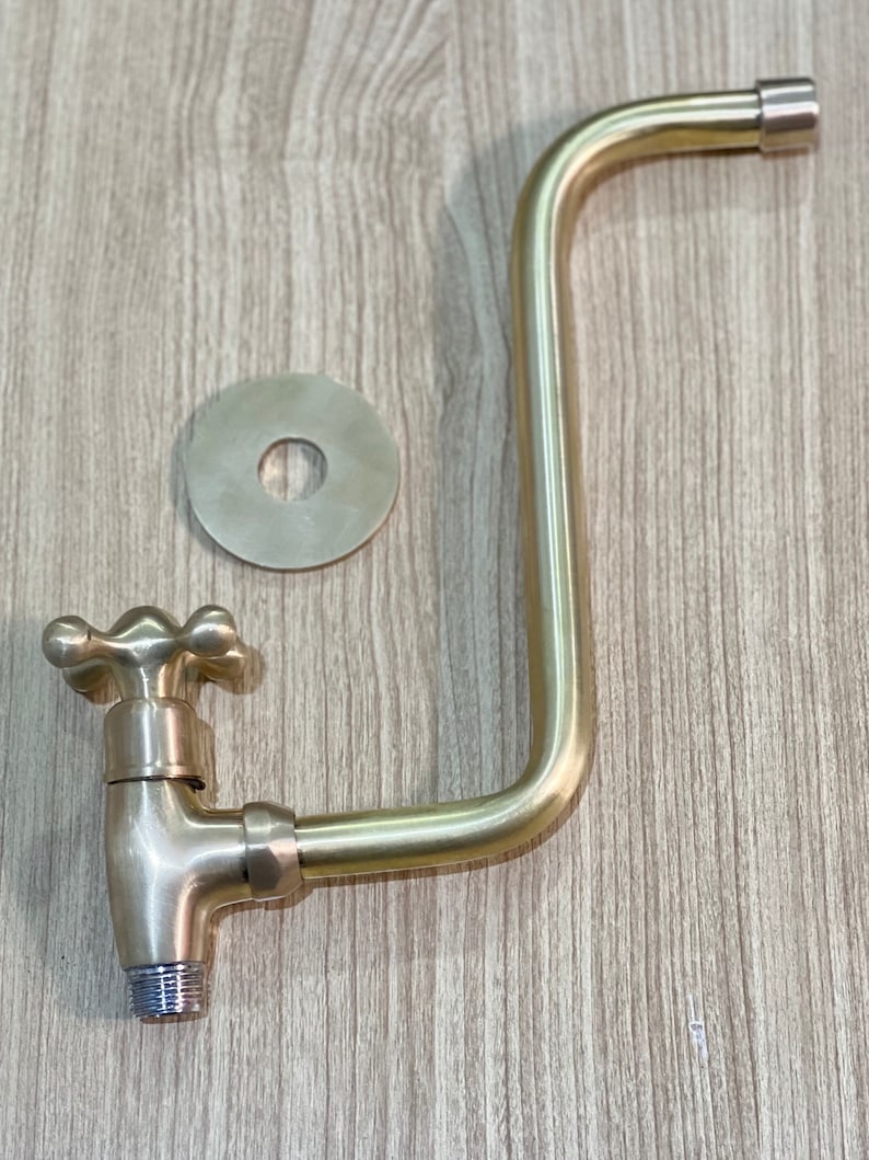 May include: A gold-toned faucet with a cross-shaped handle and a long, curved spout. The faucet is attached to a round, gold-toned wall plate.