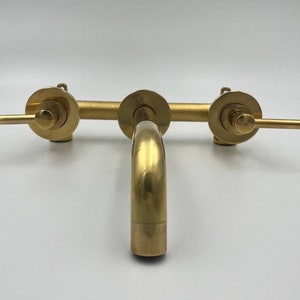 Op de afbeelding: Een goudkleurige wandgemonteerde kraan met twee handvatten en een uitloop. De kraan heeft een modern ontwerp en is gemaakt van metaal.