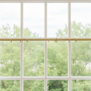 May include: A brass-coloured curtain rail with rings is mounted on a white window frame. The rail is horizontal, and the rings are evenly spaced. The window has multiple panes, with green trees visible outside.