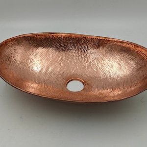 May include: Oval copper sink with a hammered texture and a central drain hole. The sink has a warm, reddish-brown color and a smooth, polished rim. This handcrafted sink is suitable for bathroom or kitchen use.