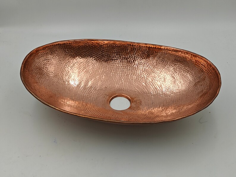 May include: Oval copper sink with a hammered texture and a central drain hole. The sink has a warm, reddish-brown color and a smooth, polished rim. This handcrafted sink is suitable for bathroom or kitchen use.