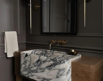 May include: A white marble sink with black veining, set into a dark wood vanity. The sink is rectangular and has a curved front edge. The vanity is made of dark wood and has a simple design.