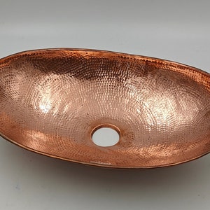 May include: Oval copper sink with a hammered texture. The sink has a polished finish and a central drain hole. This vessel sink is designed for bathroom use and adds a rustic touch.