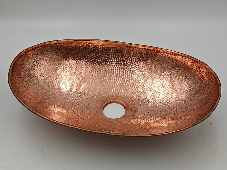 May include: Oval copper sink with a hammered texture. The sink has a polished finish and a central drain hole. This vessel sink is designed for bathroom use and adds a rustic touch.