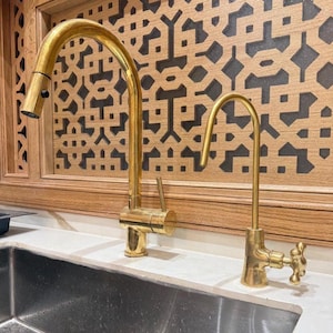 May include: Two gold-colored kitchen faucets. The larger faucet has a curved spout and a pull-down sprayer. The smaller faucet has a curved spout and a decorative handle. The faucets are installed on a white countertop with a stainless steel sink.