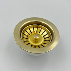 May include: A brass-colored kitchen sink strainer with a circular design. The strainer has a perforated basket and a central post. The outer rim is a lighter, silver color. The product is designed for kitchen use.