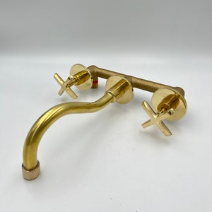 May include: A gold-toned wall-mounted faucet with two cross-shaped handles. The faucet has a curved spout and a modern design.
