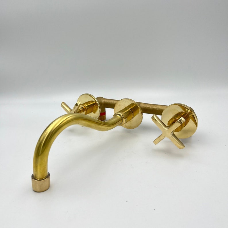 May include: A gold-toned wall-mounted faucet with two cross-shaped handles. The faucet has a curved spout and a polished finish.