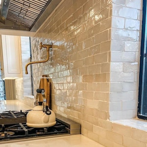 May include: A white kitchen with a gold faucet and a white kettle on a black stovetop. The kitchen has white subway tile backsplash and a window with a black frame.