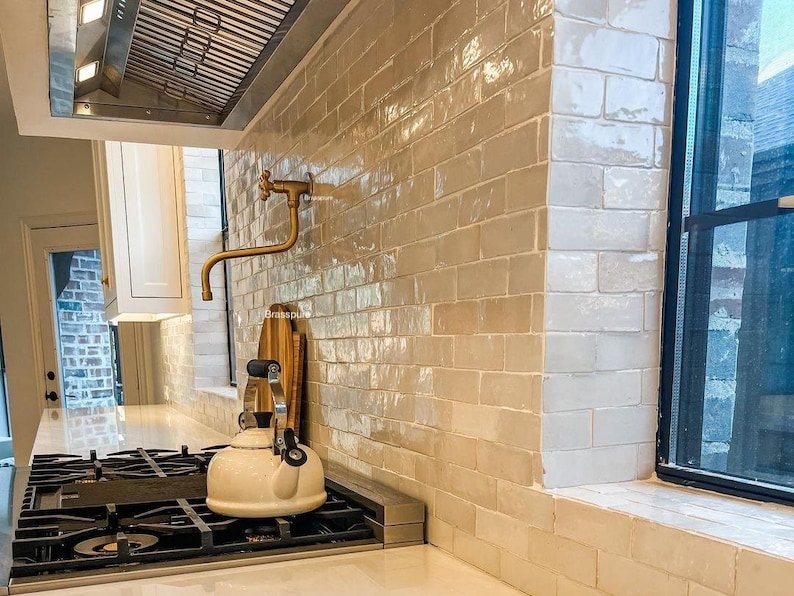 May include: A white kitchen with a gold faucet and a white kettle on a black stovetop. The kitchen has white subway tile backsplash and a window with a black frame.