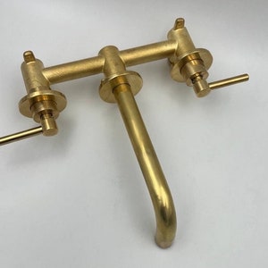 Op de afbeelding: Een gouden messing kraan met twee handvatten en een uitloop. De kraan is aan een muur gemonteerd.