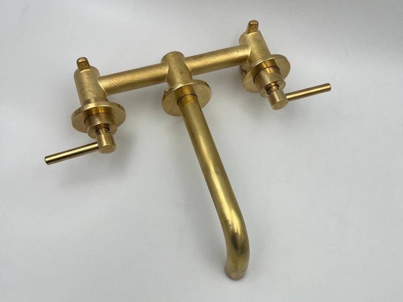 Op de afbeelding: Een gouden messing kraan met twee handvatten en een uitloop. De kraan is aan een muur gemonteerd.