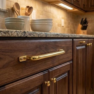 Pode incluir: Close-up de um armário de cozinha de madeira escura com uma longa maçaneta dourada. As portas do armário têm um design de painel rebaixado. Outros itens de cozinha são visíveis ao fundo, incluindo tigelas e utensílios.