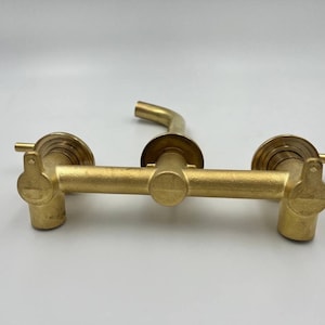 Op de afbeelding: Een wandgemonteerde kraan van verguld messing met twee handvatten en een uitloop. De kraan is ontworpen voor een badkamer- of keukenwasbak.
