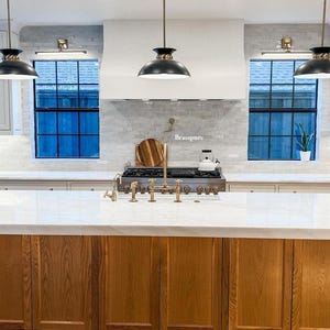 May include: A kitchen island with a white countertop and wooden cabinets. The island has a gold faucet and a stainless steel stovetop. The kitchen has white cabinets, black pendant lights, and a white subway tile backsplash. The backsplash has the word "Brasspure" written on it.