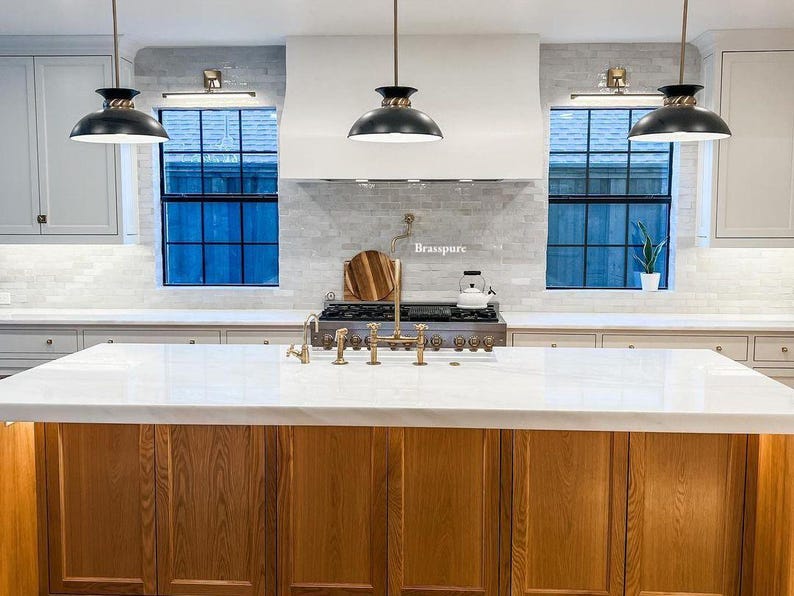 May include: A kitchen island with a white countertop and wooden cabinets. The island has a gold faucet and a stainless steel stovetop. The kitchen has white cabinets, black pendant lights, and a white subway tile backsplash. The backsplash has the word "Brasspure" written on it.