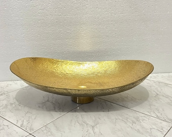 May include: A large, oval-shaped, gold-colored metal serving bowl with a hammered texture. The bowl has a wide, shallow base and a raised lip.