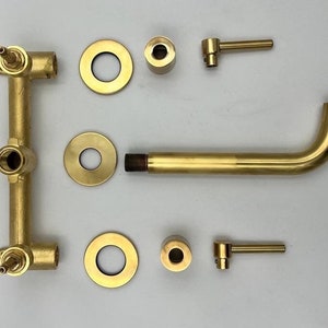 Op de afbeelding: Een gouden badkamerkraanset met twee handvatten, een uitloop en een wandmontage. De set bevat twee ronde rozetten, twee ronde knoppen en twee hendels.