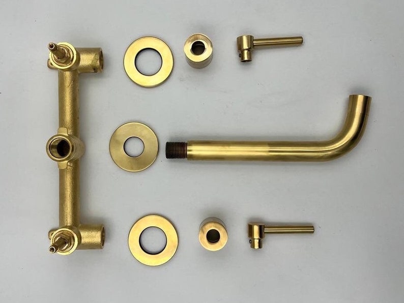 Op de afbeelding: Een gouden badkamerkraanset met twee handvatten, een uitloop en een wandmontage. De set bevat twee ronde rozetten, twee ronde knoppen en twee hendels.