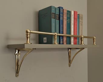 May include: A wall-mounted bookshelf with a gray shelf, brass railing, and brass brackets. Several books with various colored spines are displayed on the shelf, creating a decorative and functional storage solution.