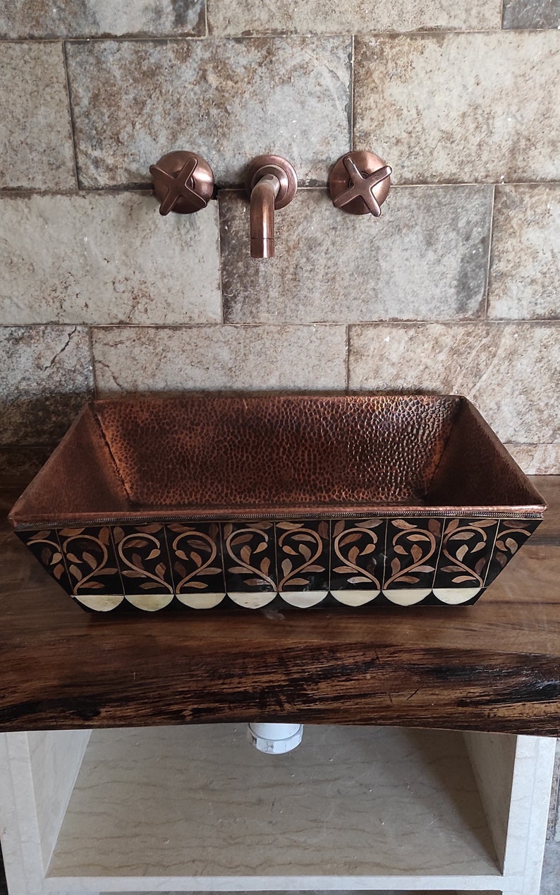 K&ouml;nnte beinhalten: Ein Kupferwaschbecken mit geh&auml;mmerter Oberfl&auml;che und einem dekorativen Schwarz-Wei&szlig;-Design. Das Waschbecken ist auf einer Holzarbeitsplatte mit einem wei&szlig;en Regal darunter montiert.