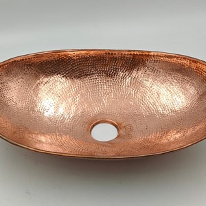 May include: Oval copper sink with a hammered texture and a drain hole. The sink has a polished finish, reflecting light. The copper color gives it a warm, rustic appearance. This vessel sink is designed for bathroom use.