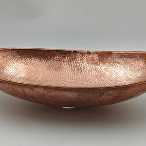 May include: A hammered copper oval bowl with a textured surface. The bowl has a wide, shallow design and a rich, reddish-brown color. The interior and exterior of the bowl are covered in a pattern of small, evenly spaced indentations.