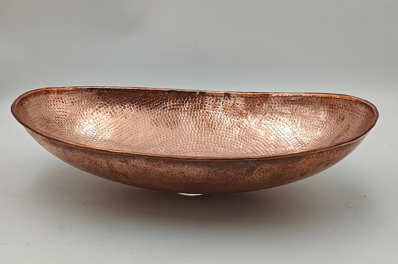 May include: A hammered copper oval bowl with a textured surface. The bowl has a wide, shallow design and a rich, reddish-brown color. The interior and exterior of the bowl are covered in a pattern of small, evenly spaced indentations.