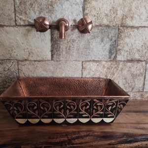 K&ouml;nnte beinhalten: Ein geh&auml;mmertes Kupferwaschbecken mit einem dekorativen schwarzen und wei&szlig;en Blumendesign. Das Waschbecken ist auf einer Holzarbeitsplatte mit Kupferarmaturen montiert.