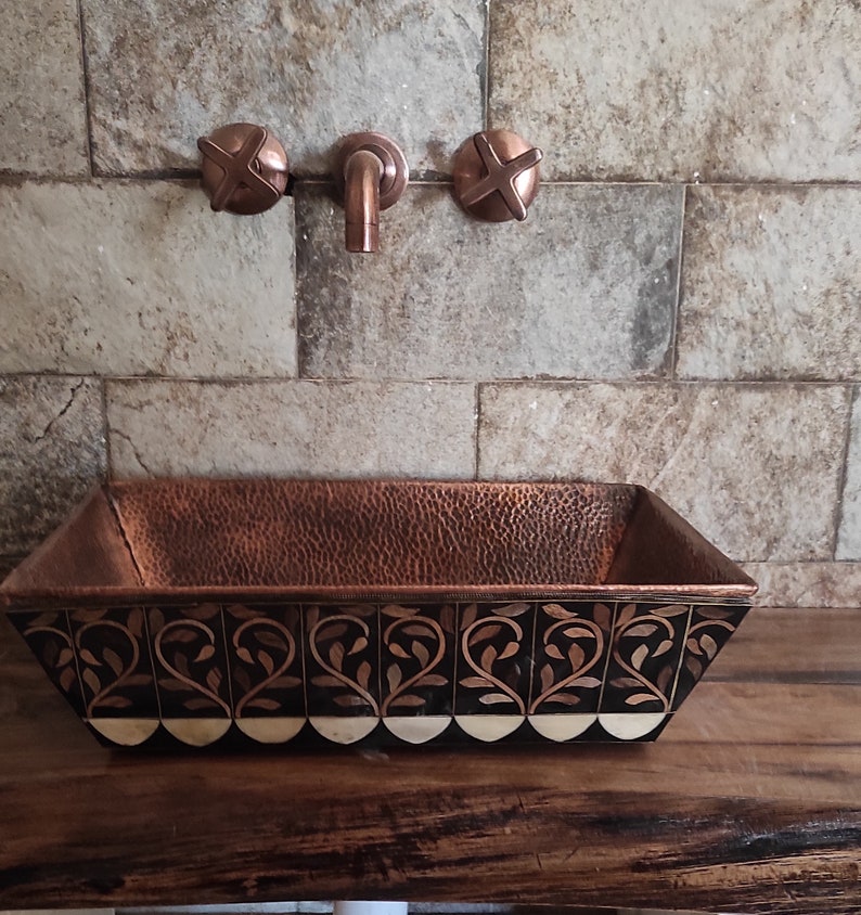 K&ouml;nnte beinhalten: Ein geh&auml;mmertes Kupferwaschbecken mit einem dekorativen schwarzen und wei&szlig;en Blumendesign. Das Waschbecken ist auf einer Holzarbeitsplatte mit Kupferarmaturen montiert.