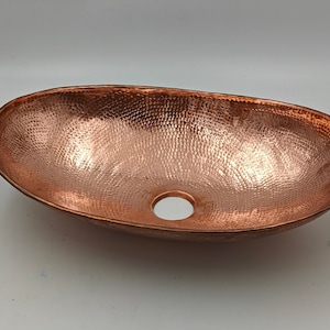 May include: Oval copper sink with a hammered texture. The sink has a polished finish and a drain hole. This vessel sink is a bathroom fixture.