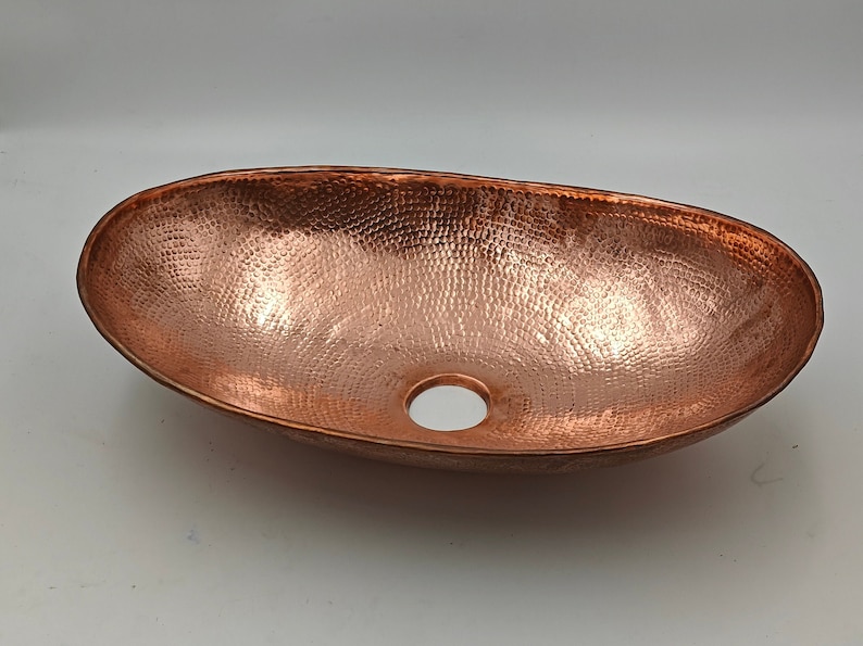 May include: Oval copper sink with a hammered texture. The sink has a polished finish and a drain hole. This vessel sink is a bathroom fixture.