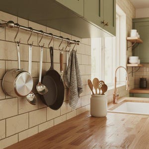May include: A stainless steel wall-mounted rail with S-hooks holding a silver saucepan, two silver ladles, a black cast iron skillet, a wooden-handled tool, and a grey patterned kitchen towel against a beige subway tile backsplash.