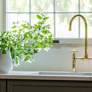 May include: A white ceramic jug filled with a bouquet of white flowers with green leaves sits on a white countertop next to a gold kitchen tap with two handles.