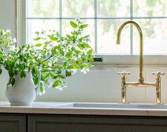 May include: A white ceramic pitcher filled with a bouquet of white flowers with green leaves sits on a white countertop next to a gold kitchen faucet with two handles.