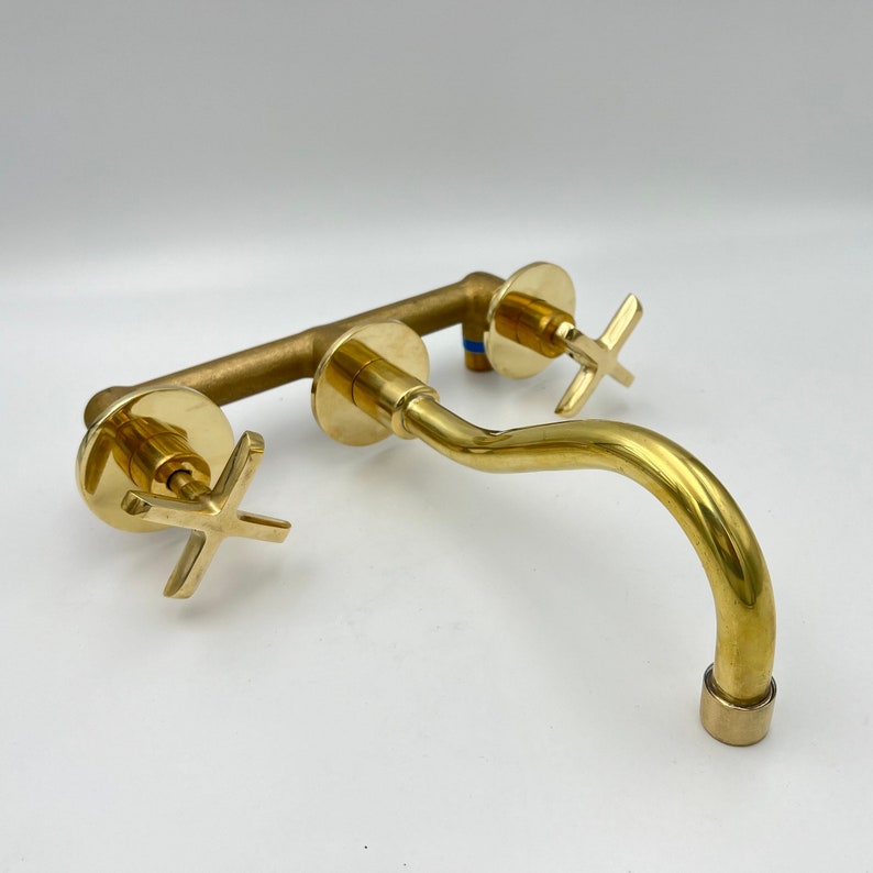 May include: A gold-toned wall-mounted bathroom faucet with two cross-shaped handles and a curved spout.