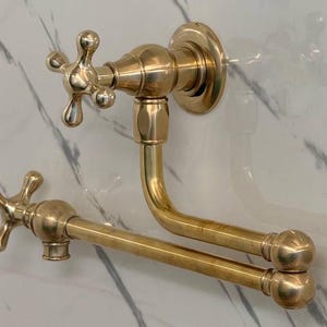 May include: A gold-toned kitchen tap with a swivel spout and two cross-shaped handles. The tap is mounted to a wall.