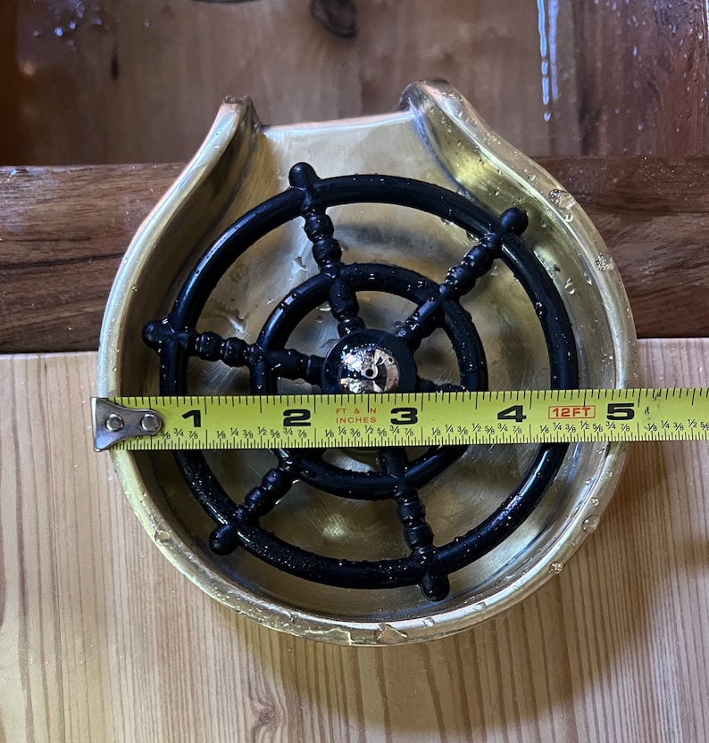 May include: A brass soap dish with a black plastic ship's wheel design. The dish is 4 inches in diameter.
