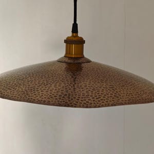 Könnte beinhalten: Kupferfarbene Pendelleuchte mit gehämmert strukturierter Schirm. Der Schirm ist breit und flach, mit einer messingfarbenen Halterung und einem schwarzen Kabel. Das Licht hängt von der Decke.
